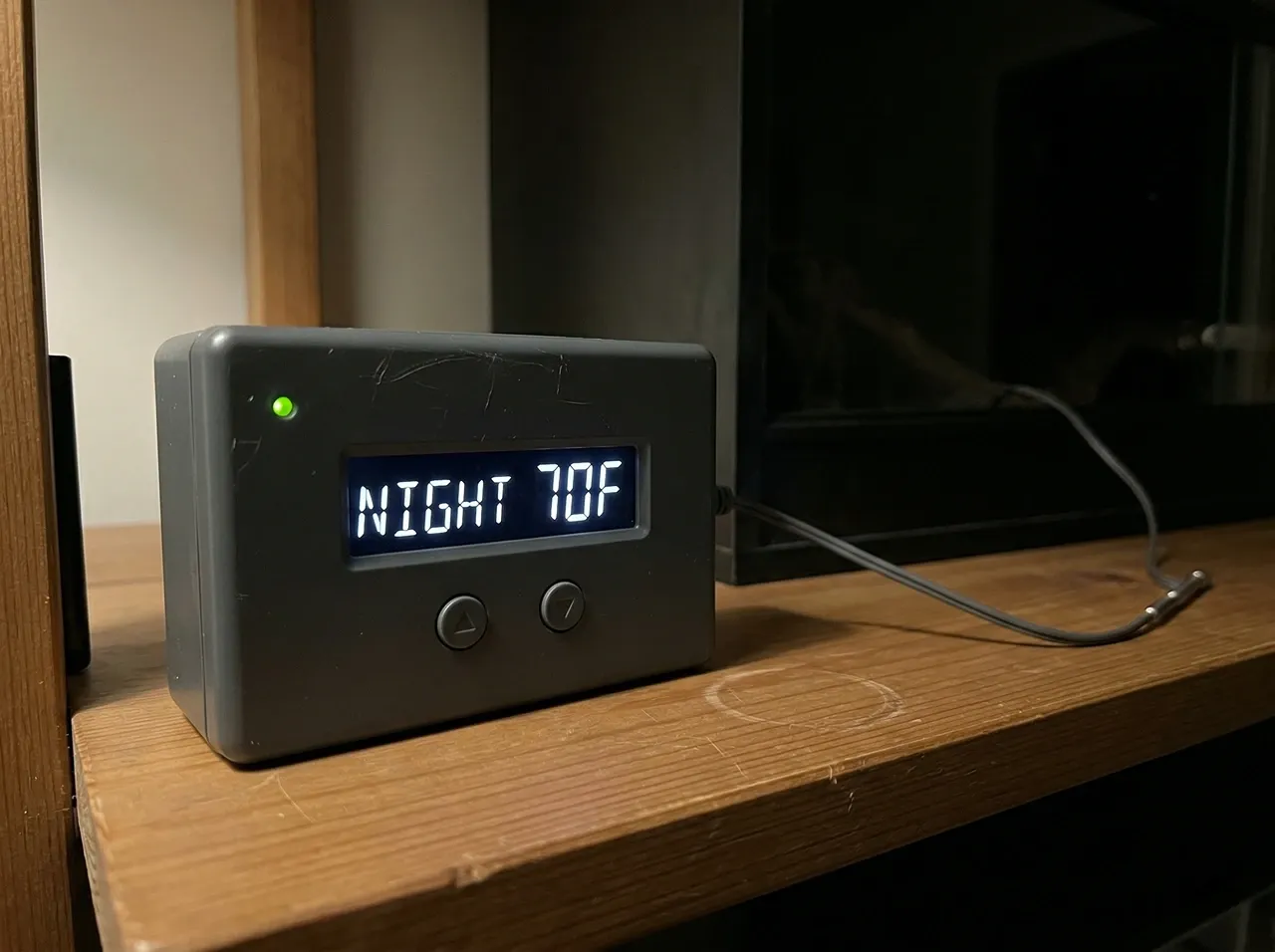 Reptile thermostat displaying NIGHT 70F on a wooden shelf beside a dark bearded dragon enclosure
